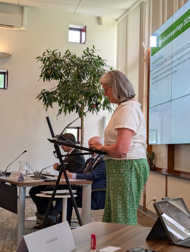 Winny Toersen staat met wit shirt en groene broek achter het katheder voor de raadszaal.
