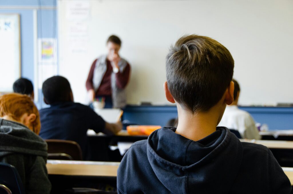 Kind in schoolklas vanaf achter gefotografeerd. Op achtergrond de leerkracht leunend tegen de muur.