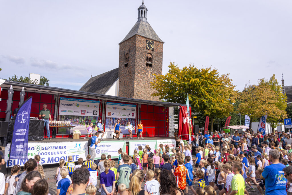 Kinderen en volwassenen in sportkleding doen mee met de warming-up die op de platte kar wordt gedemonstreerd.