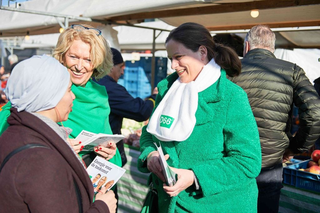 Drie vrouwen in gesprek op een markt, twee gekleed in groene D66-kleding, lachend en brochures uitdelend over D66 en politieke betrokkenheid. De sfeer is open en vriendelijk, met een achtergrond van marktkramen en mensen – een uitnodigend beeld dat vrouwen aanmoedigt om politiek actief te worden bij D66 Hoeksche Waard.